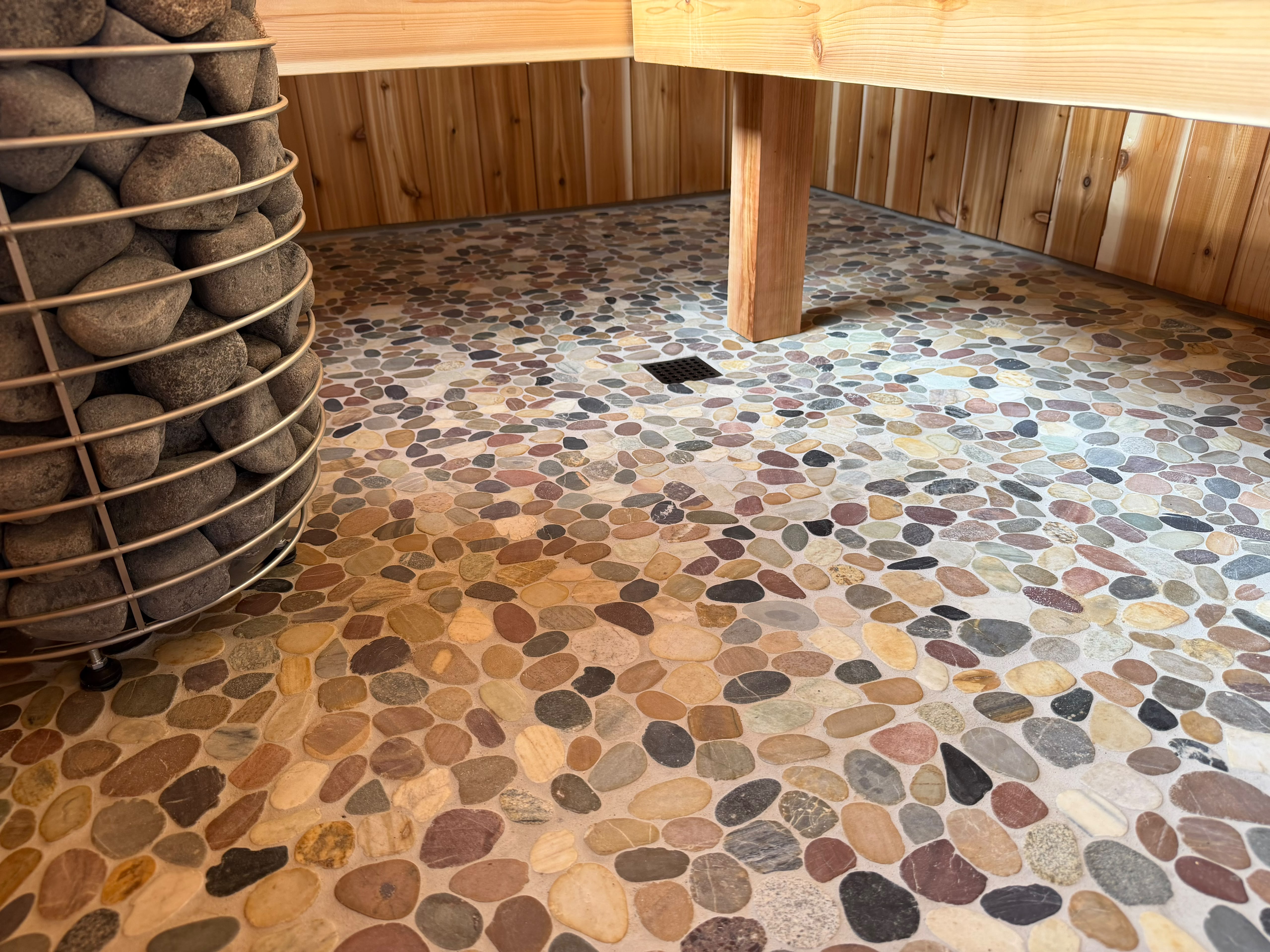 Pebble-mosaic sauna floor with a stone-filled heater basket and cedar bench